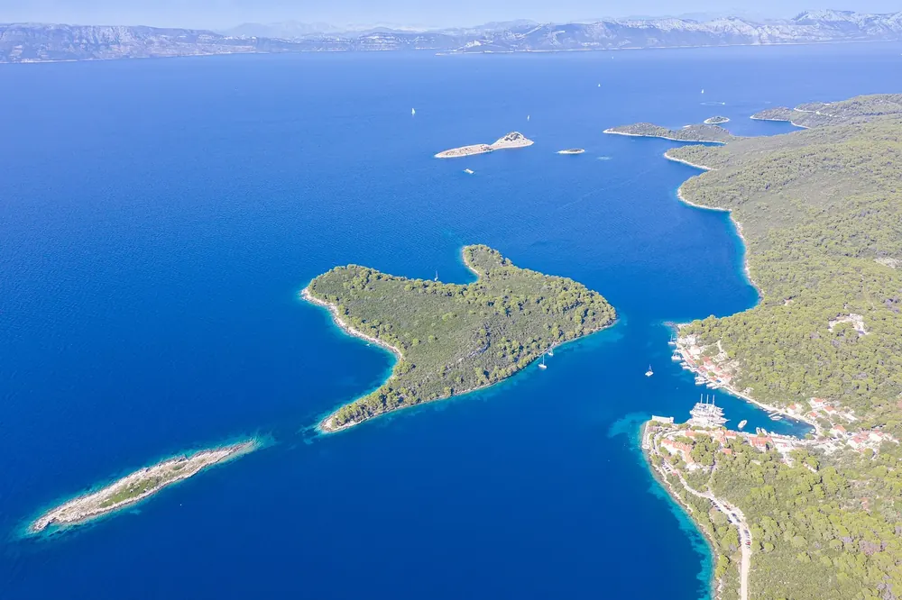 Aerial view of Pomena on Mljet, the start of Route A into Mljet National Park: a small harbour village on a bay, with the wooded islet of Pomeštak in the deep blue sea