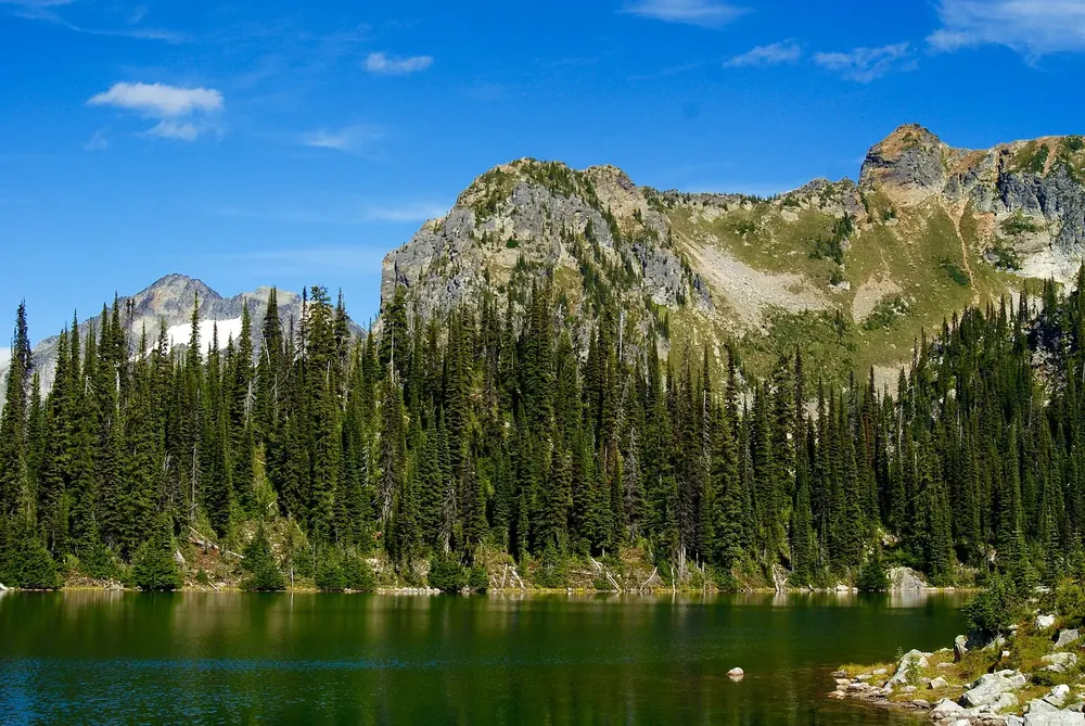 Eva Lake in Mount Revelstoke National Park: a deep blue mountain lake in front of dense conifer forest and rocky peaks under a blue sky.