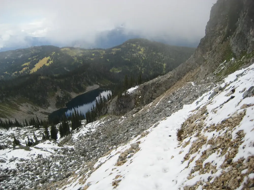 View from the Jade Pass trail in Mount Revelstoke National Park down to the dark Miller Lake among snowfields, scree and conifer forest.