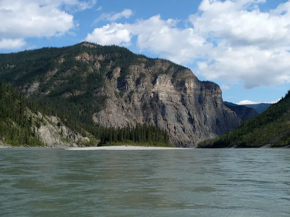 Third Canyon on the South Nahanni River in Nahanni National Park Reserve: a wide river in front of steep, forested limestone walls under clouds