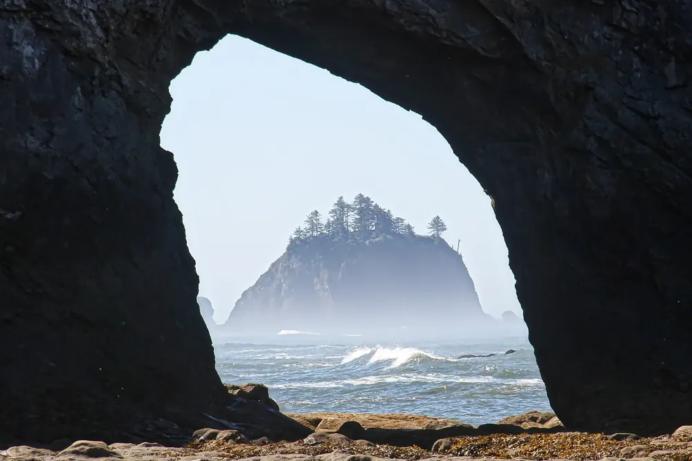 Hole-in-the-Wall in Olympic National Park: looking through a dark rock arch on the beach to a forested rock island in the sea haze