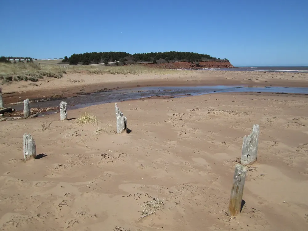 North Rustico Beach in Prince Edward Island National Park: red sand beach with old wooden posts and a lagoon, a wooded headland behind.