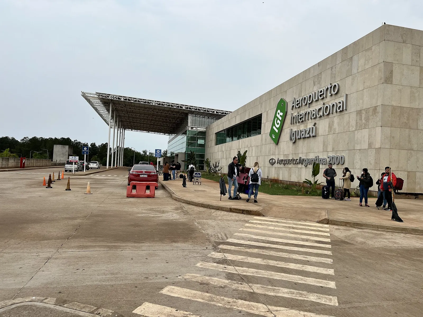 Puerto Iguazú airport: terminal with a stone facade and the lettering “Aeropuerto Internacional Iguazú”, a zebra crossing and travellers with luggage in front
