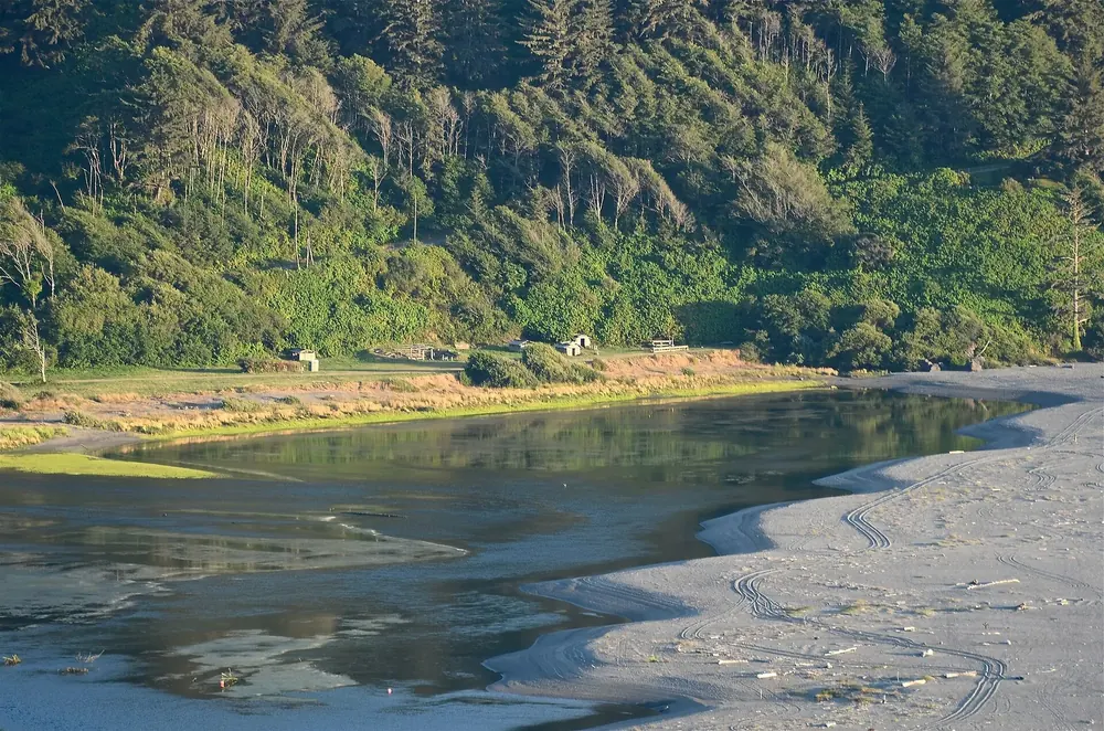 Mouth of the Klamath River in Redwood National Park: calm water behind a sandbar, wooden huts of the Yurok Tribe on the shore in front of a forested slope
