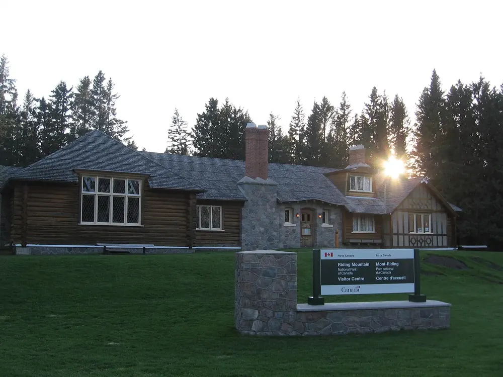 Visitor centre of Riding Mountain National Park in Wasagaming: a log building with a stone chimney, lawn and sign in front