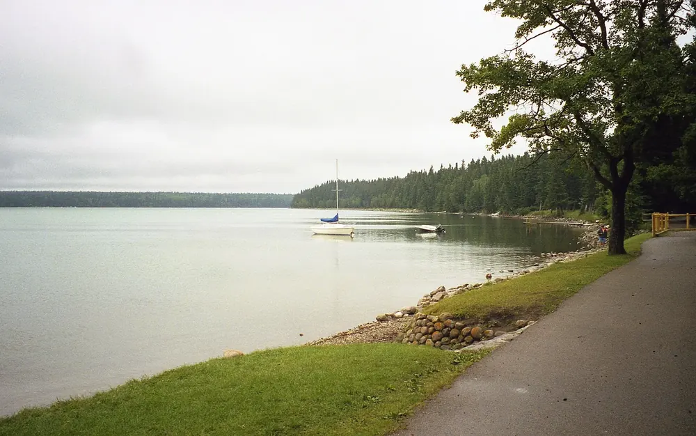 Lakeshore in Wasagaming in Riding Mountain National Park: shoreline path with lawn and stone edging, a sailing boat in front of a forested shore