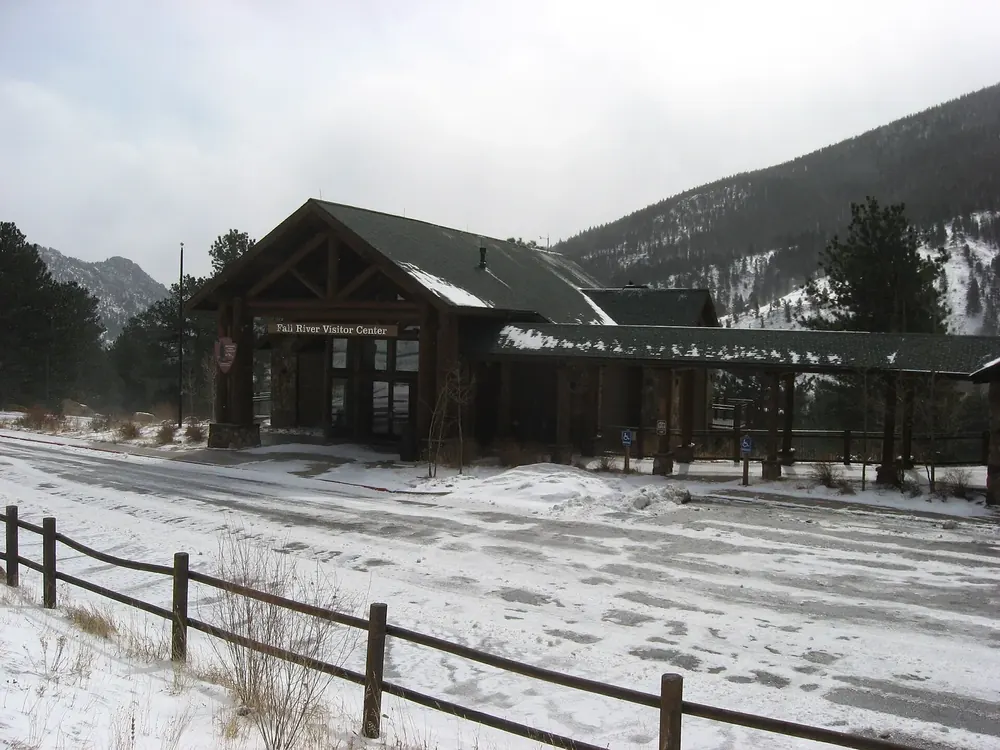 Fall River Visitor Center in winter: timber building with steep roofs, a snow-covered square and fence in front, mountains behind