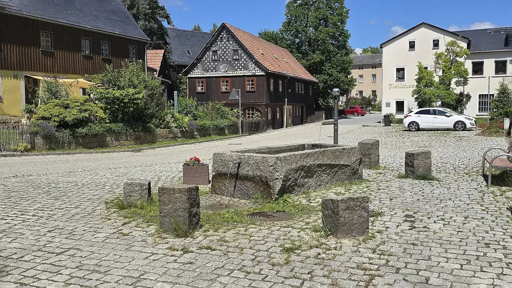 Village square in Hinterhermsdorf: a stone trough fountain on cobblestones, half-timbered Umgebinde houses with slate facades behind