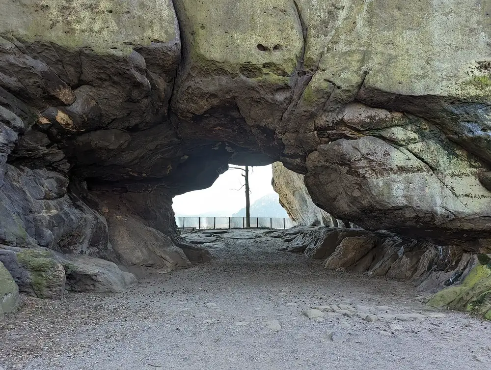 The Kuhstall rock arch on the Neuer Wildenstein in Saxon Switzerland National Park: a large hollow in the sandstone with a railing and forest beyond