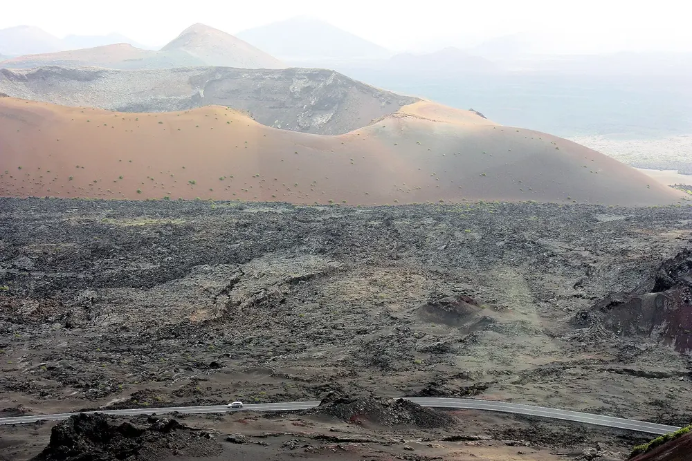 View across a dark lava field in Timanfaya National Park to the pale, wide Caldera del Corazoncillo, with the LZ-67 road at the edge