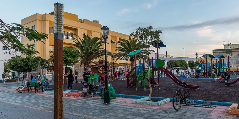 Square in Corralejo with a playground, palm trees and street lamps in front of colourful houses in the evening light