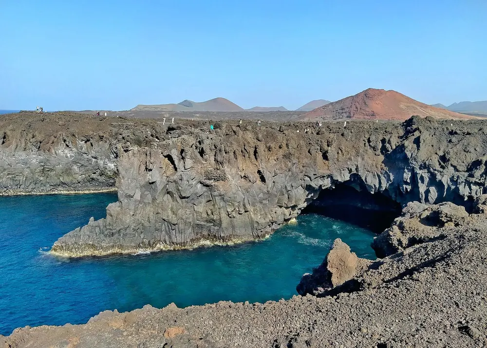 Los Hervideros: rugged lava cliffs with caves above a deep blue sea, red volcanic cones behind