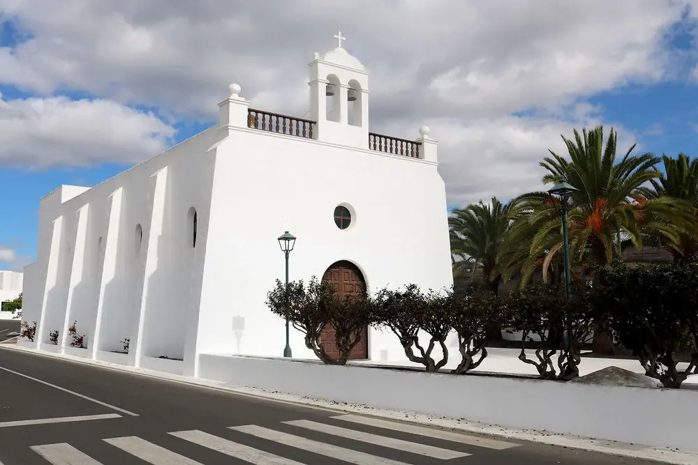 Iglesia San Isidro Labrador in Uga: white church with a bell gable and wooden doorway among palm trees