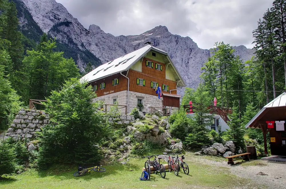 Aljažev dom mountain hut in the Vrata Valley: a wooden house with a steep roof below the massive Triglav North Face, bicycles in front