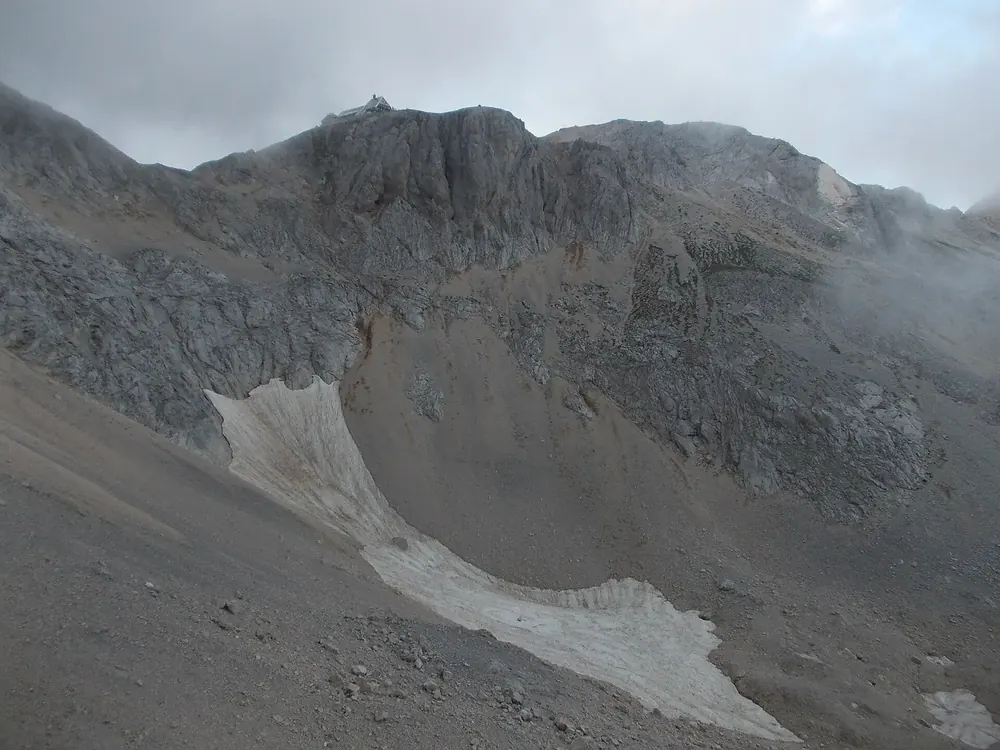 Triglavski dom na Kredarici: mountain hut on a bare ridge below grey rock faces, patches of snow on the slope