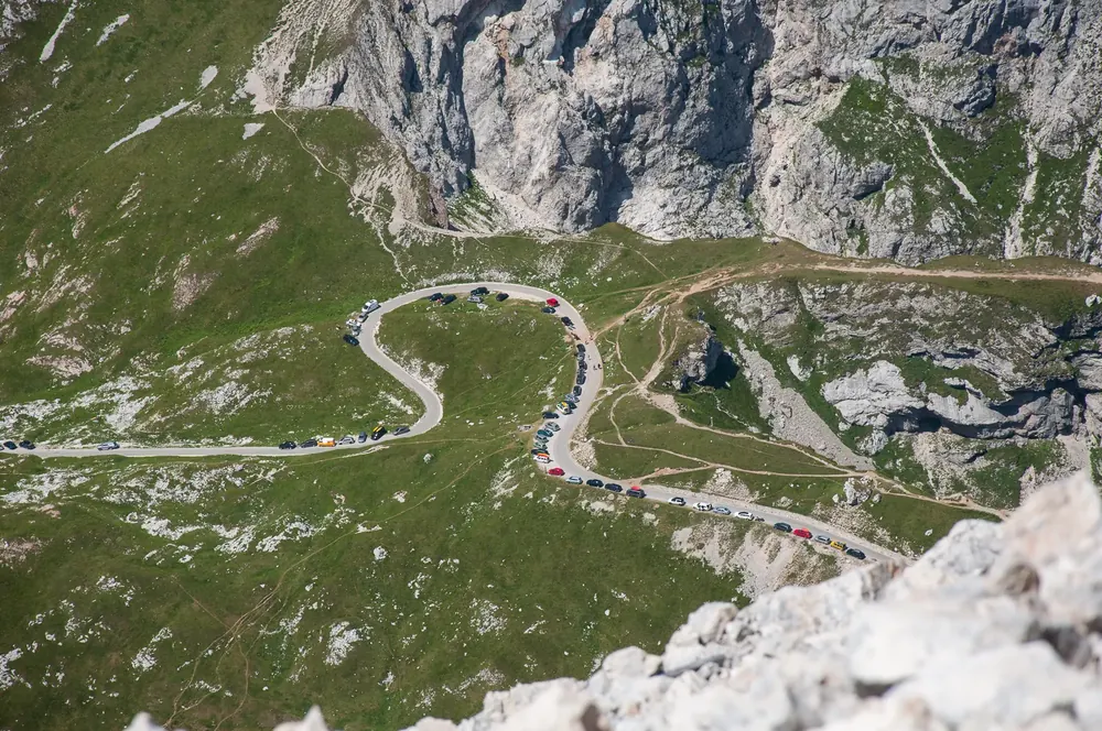 Hairpin bends of the Mangart Road in Triglav National Park on green alpine slopes below the limestone walls of Mangart, lined with cars
