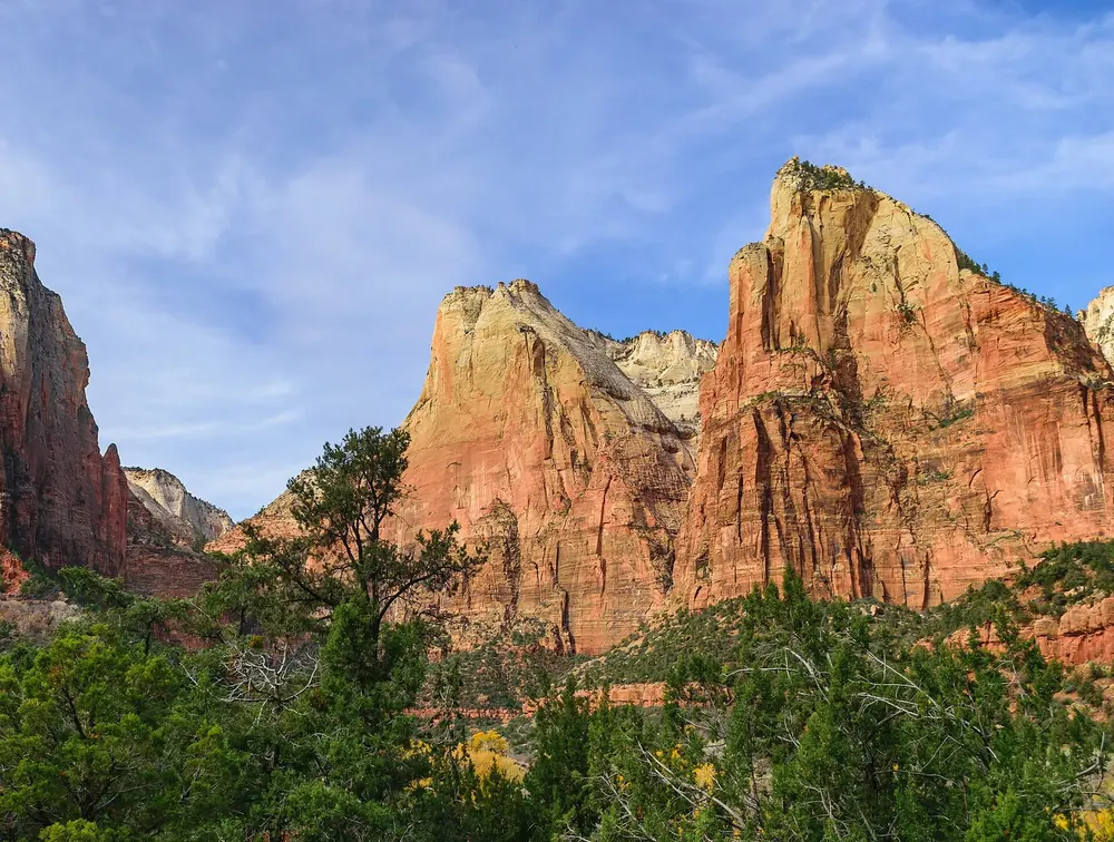 Court of the Patriarchs in Zion National Park: three massive reddish-white sandstone peaks above green deciduous woodland under a blue sky