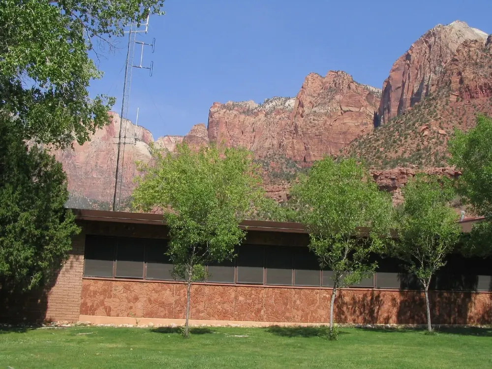 Zion Human History Museum: a low brick building among young trees, with the red rock walls of Zion Canyon behind