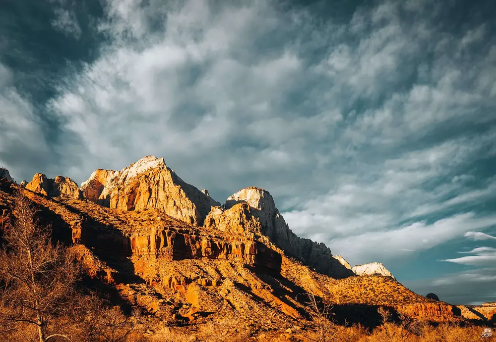 The Watchman in Zion National Park in golden evening light: a jagged sandstone peak above reddish rock ledges and a dramatic cloudy sky.