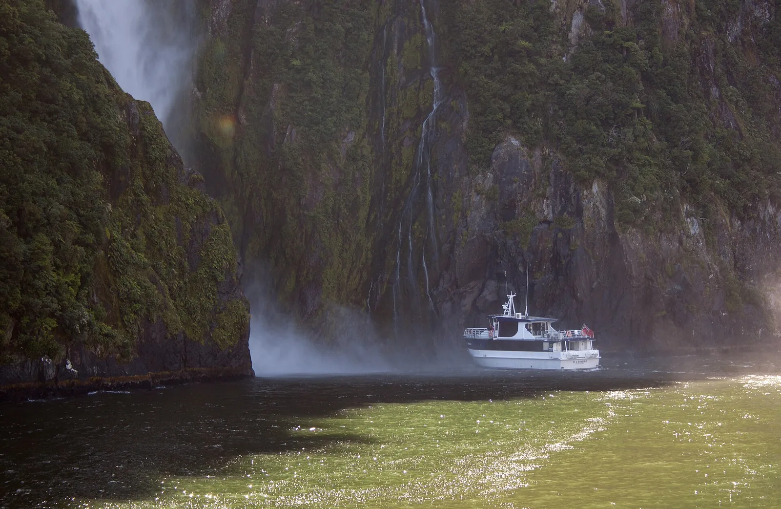 A sightseeing boat on Milford Sound heads into the spray of Stirling Falls below a steep, forested rock wall