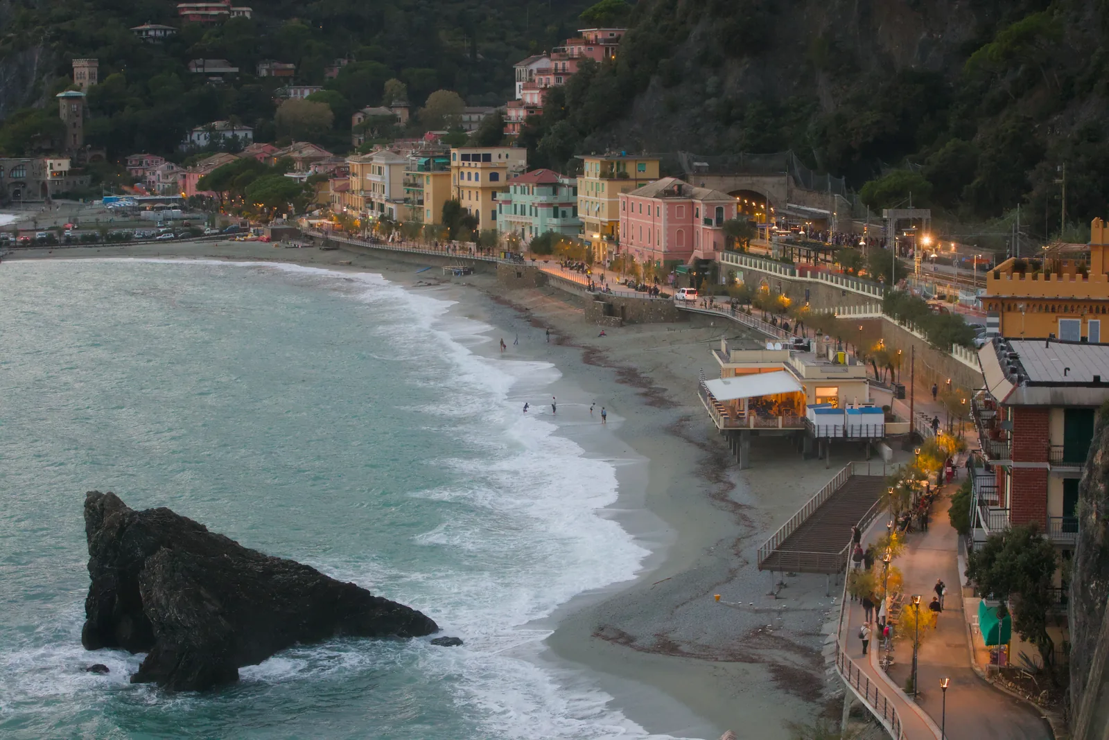 Monterosso al Mare at dusk: the beach of Fegina with a rock in the surf and the lit promenade behind