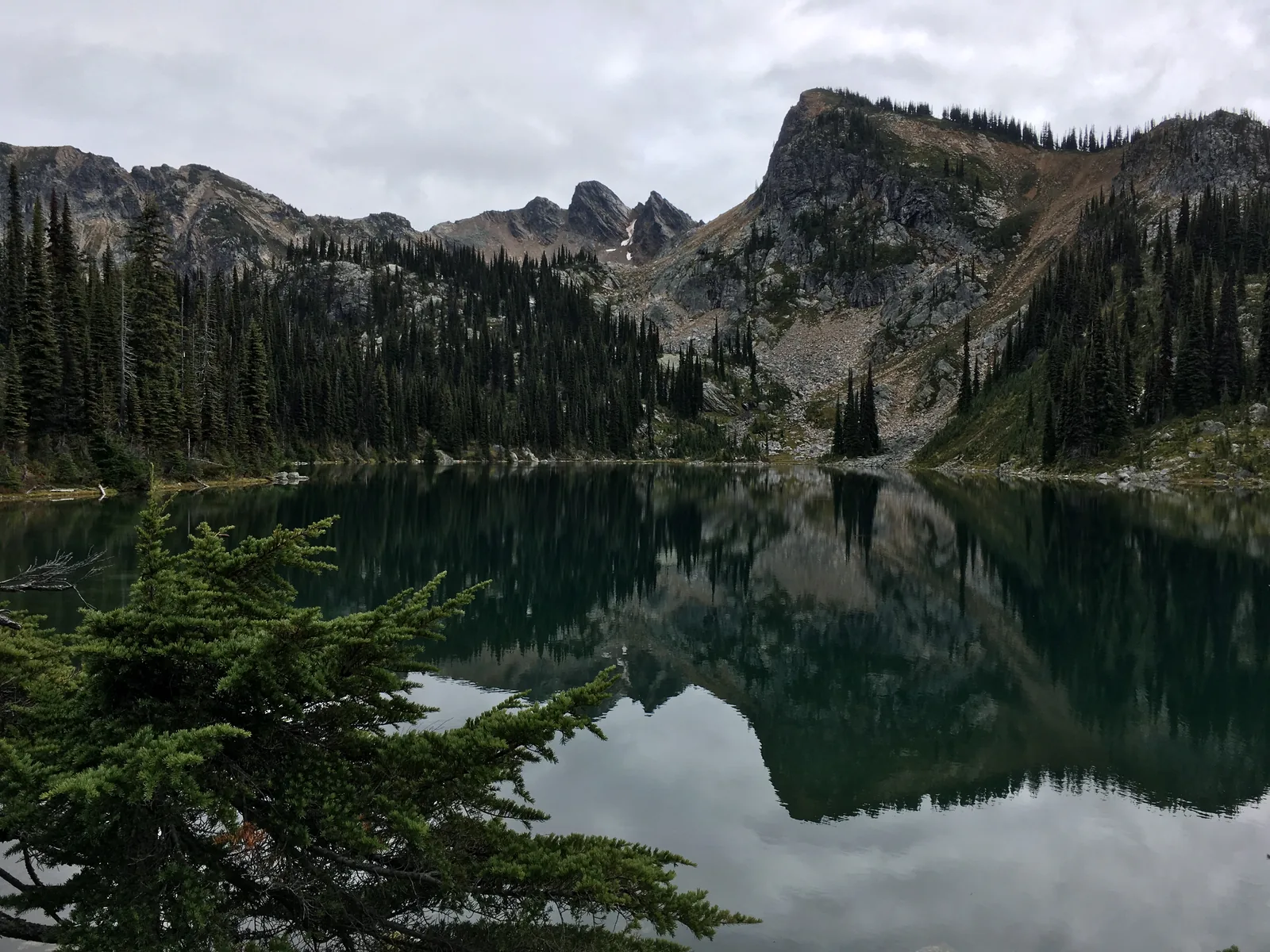 The still Eva Lake in Mount Revelstoke National Park reflects a jagged, partly snow-covered rock wall, with scattered conifers on the shore.