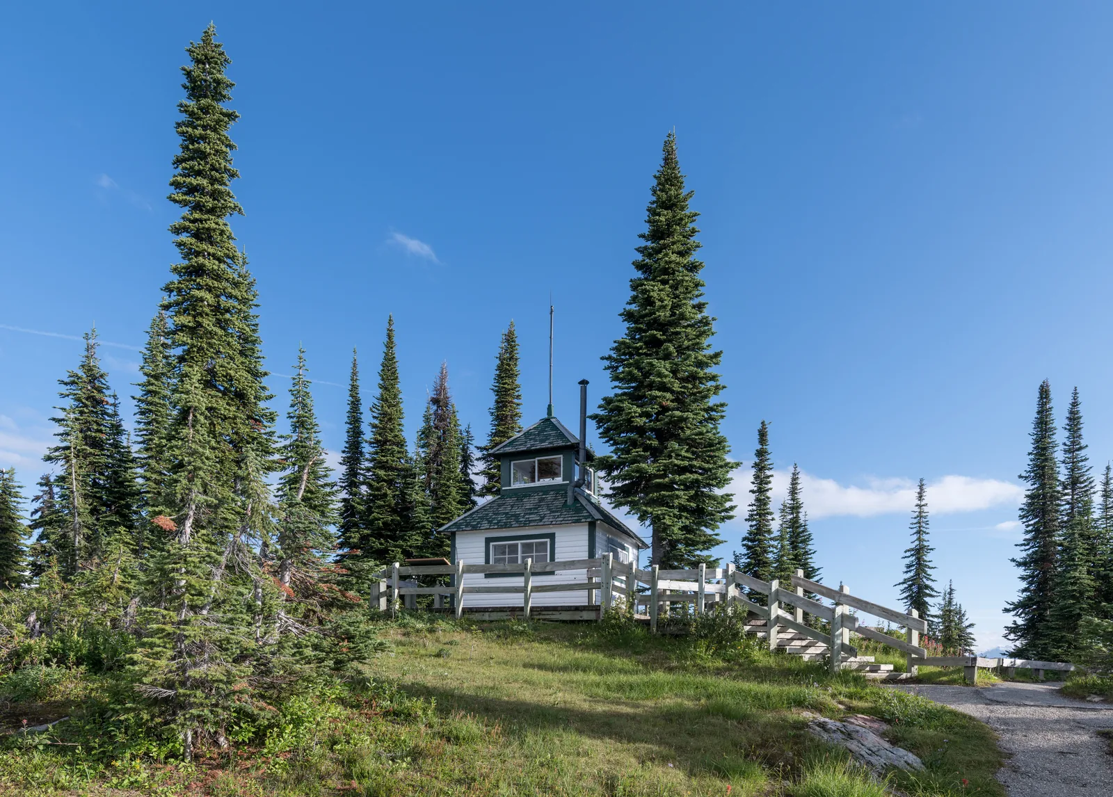 A white wooden fire lookout building stands on the summit meadow in Mount Revelstoke National Park among scattered conifers, with a wooden fence and a path in front.