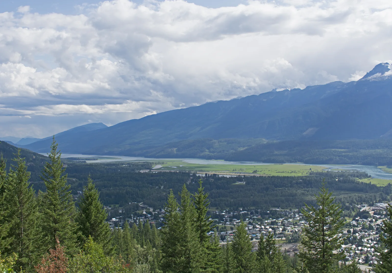 View from the mountain road in Mount Revelstoke National Park over forested slopes to the town of Revelstoke and the wide, silvery band of the Columbia River, with mountain ranges behind.