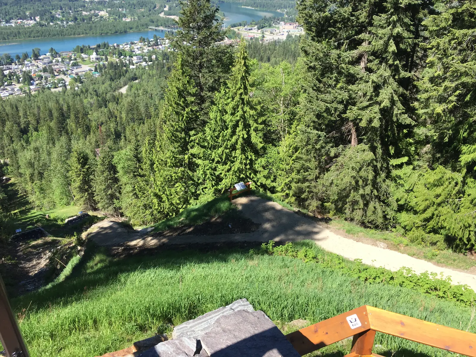 View from the top of the old ski jump in Mount Revelstoke National Park down the overgrown slope to the town of Revelstoke and the river, with a wooden railing in the foreground