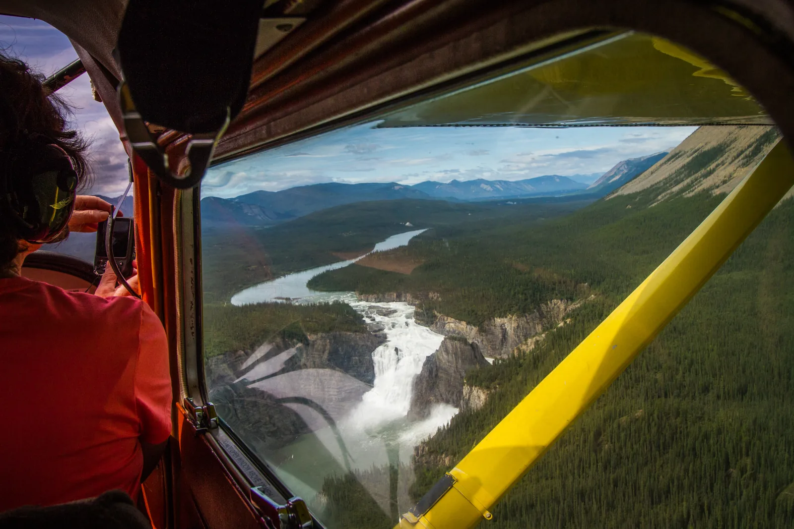 View from the window of a small aircraft in Nahanni National Park Reserve over Náįlįcho (Virginia Falls) and the South Nahanni River, with the pilot and wing strut in the foreground