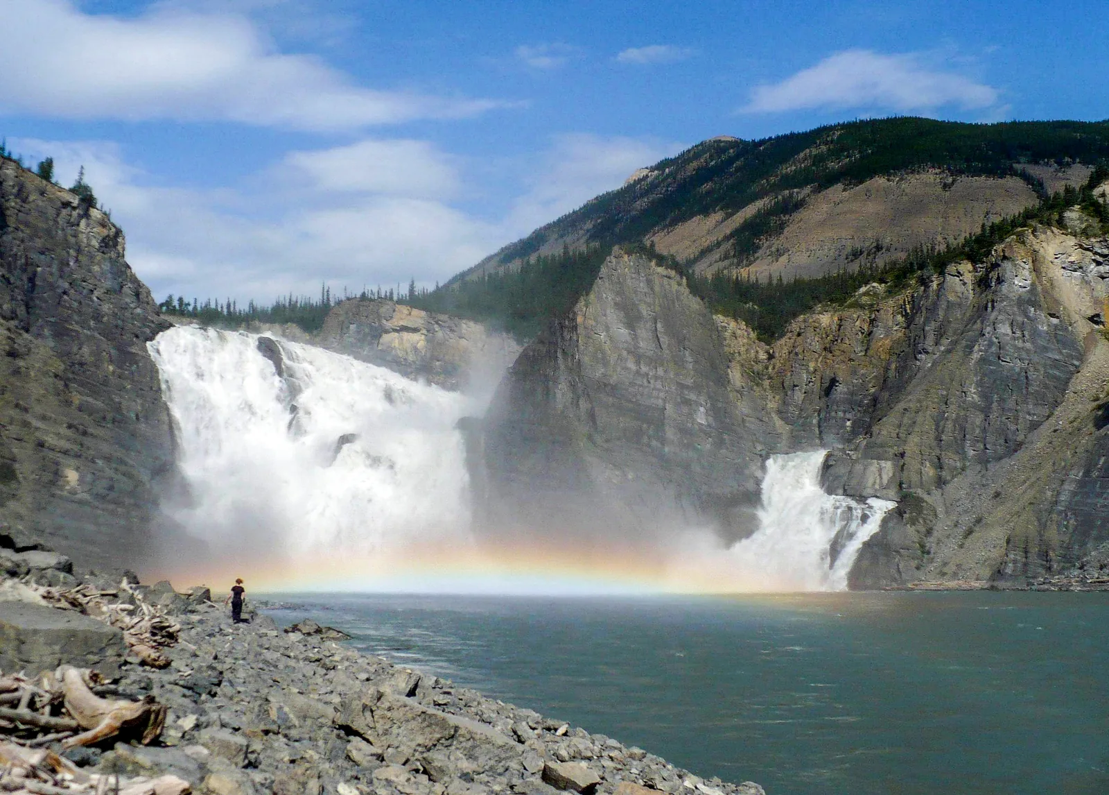 Náįlįcho (Virginia Falls) in Nahanni National Park Reserve seen from the shore: two mighty arms of the waterfall plunge into a gorge, a rainbow hangs in the spray, a tiny figure stands on the bank