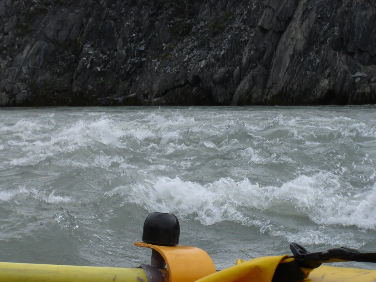 Whitewater on the Nahʔą Dehé in Nahanni National Park Reserve from a raft: foaming waves in front of a dark rock wall, a helmet in the foreground