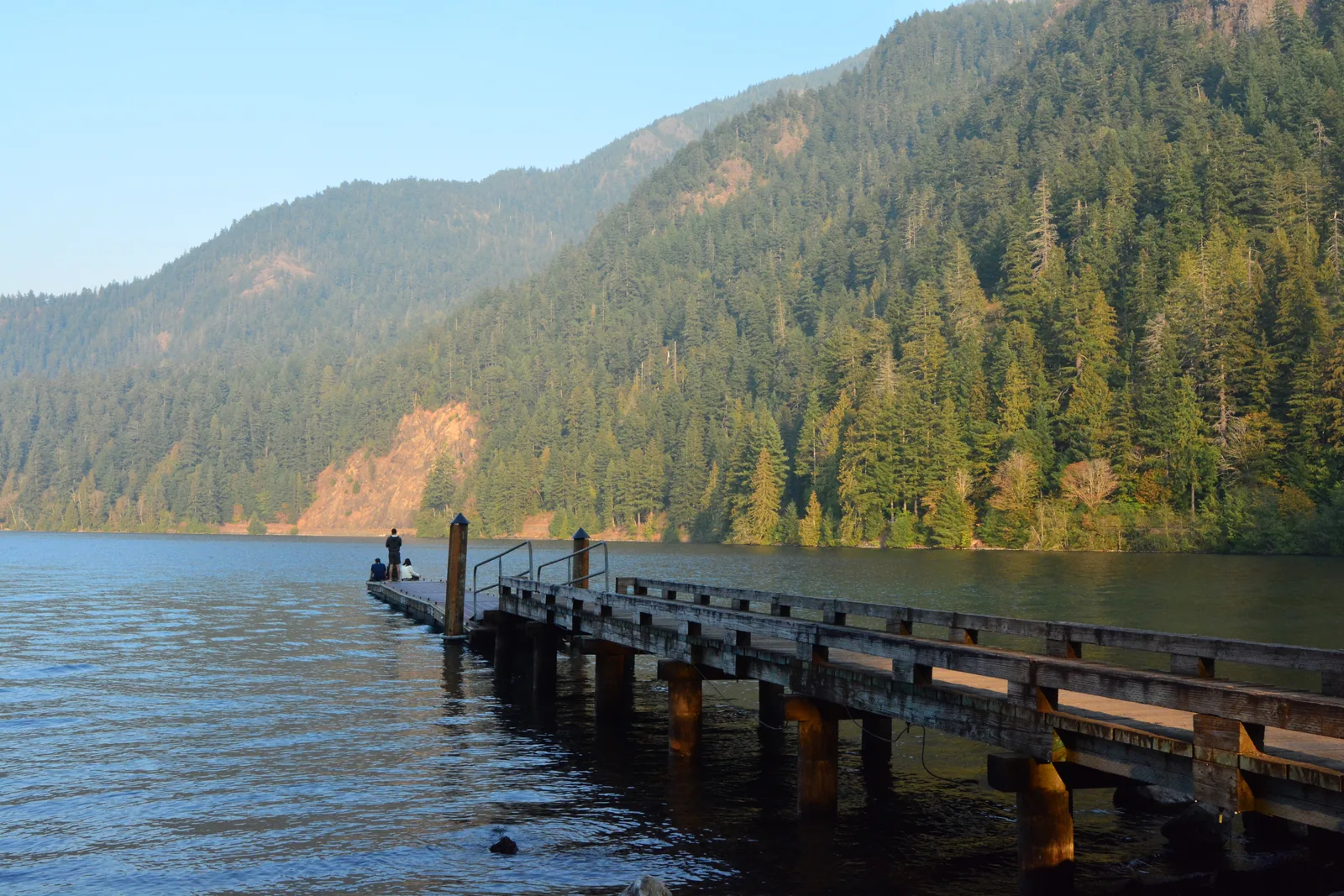 A wooden jetty reaching into the still water of Lake Crescent in Olympic National Park, with forested slopes in the evening light behind