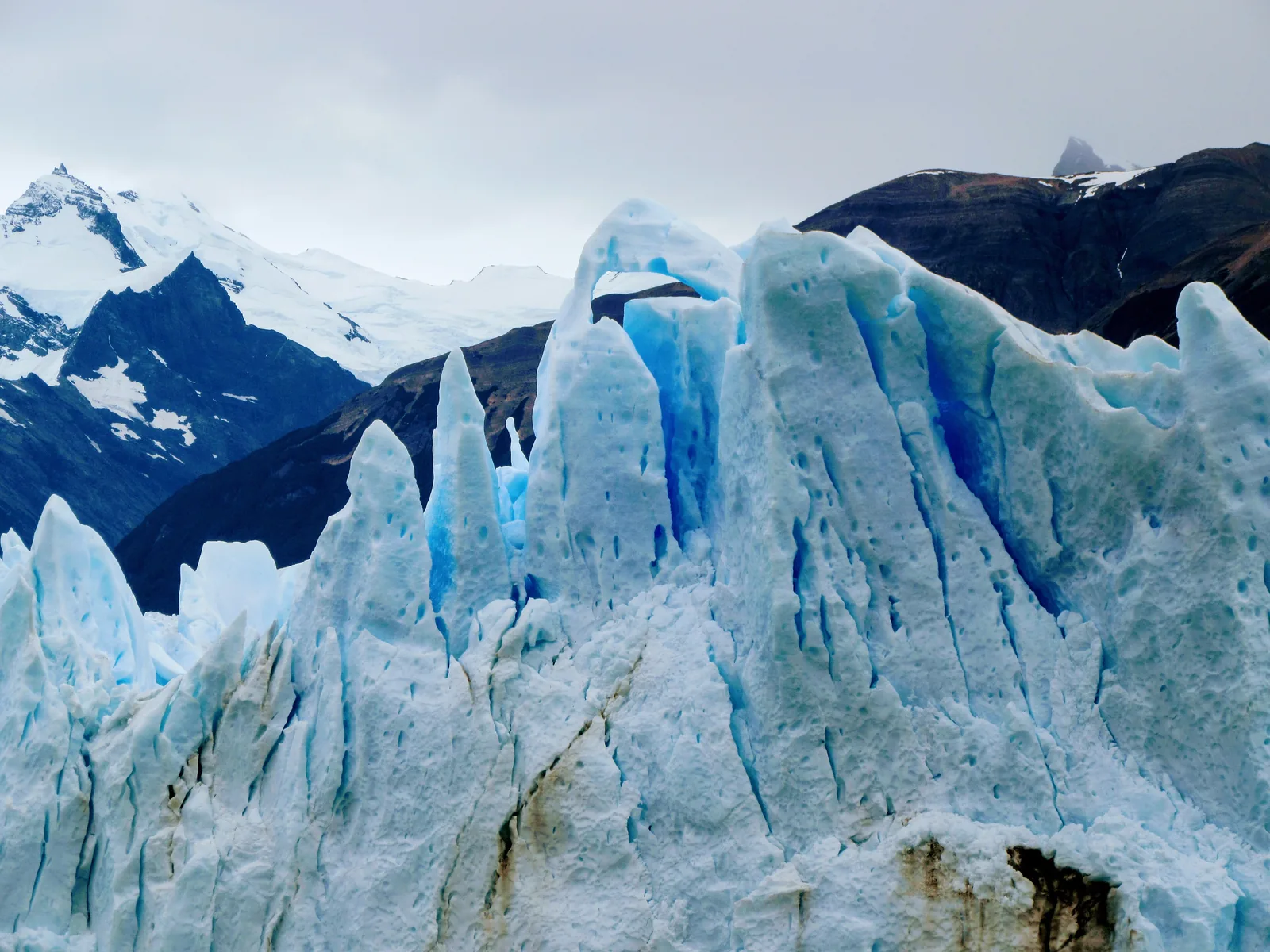 Blue ice spikes and an arch in the front of the Perito Moreno Glacier in Los Glaciares National Park, dark mountains behind