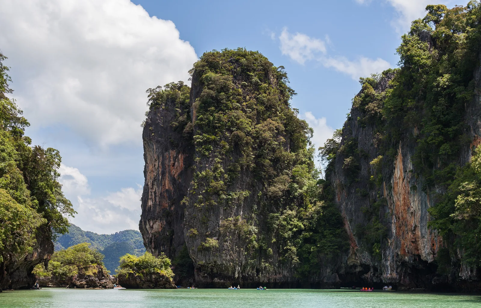 Steep, overgrown limestone cliffs of Ko Panak in Ao Phang Nga National Park, with shallow green water and a small rock islet in front