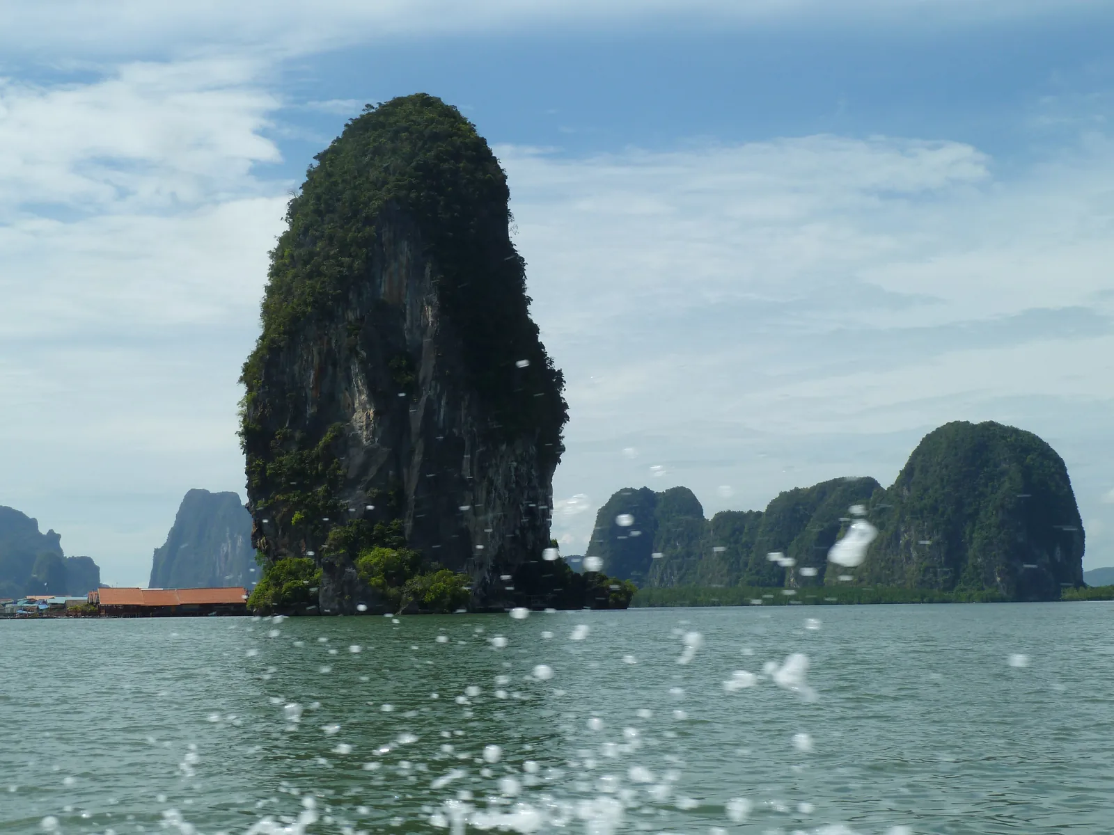 A tall overgrown limestone rock off Ko Panyi in Ao Phang Nga National Park, with the roofs of the stilt village by the water on the left