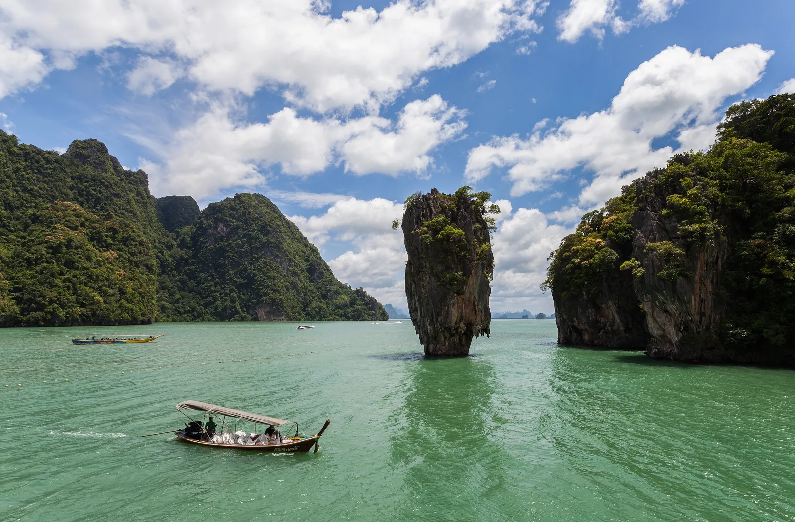 The narrow limestone pinnacle of Ko Tapu rising from green water in Ao Phang Nga National Park, the cliff of Khao Phing Kan on the right, a longtail boat in front