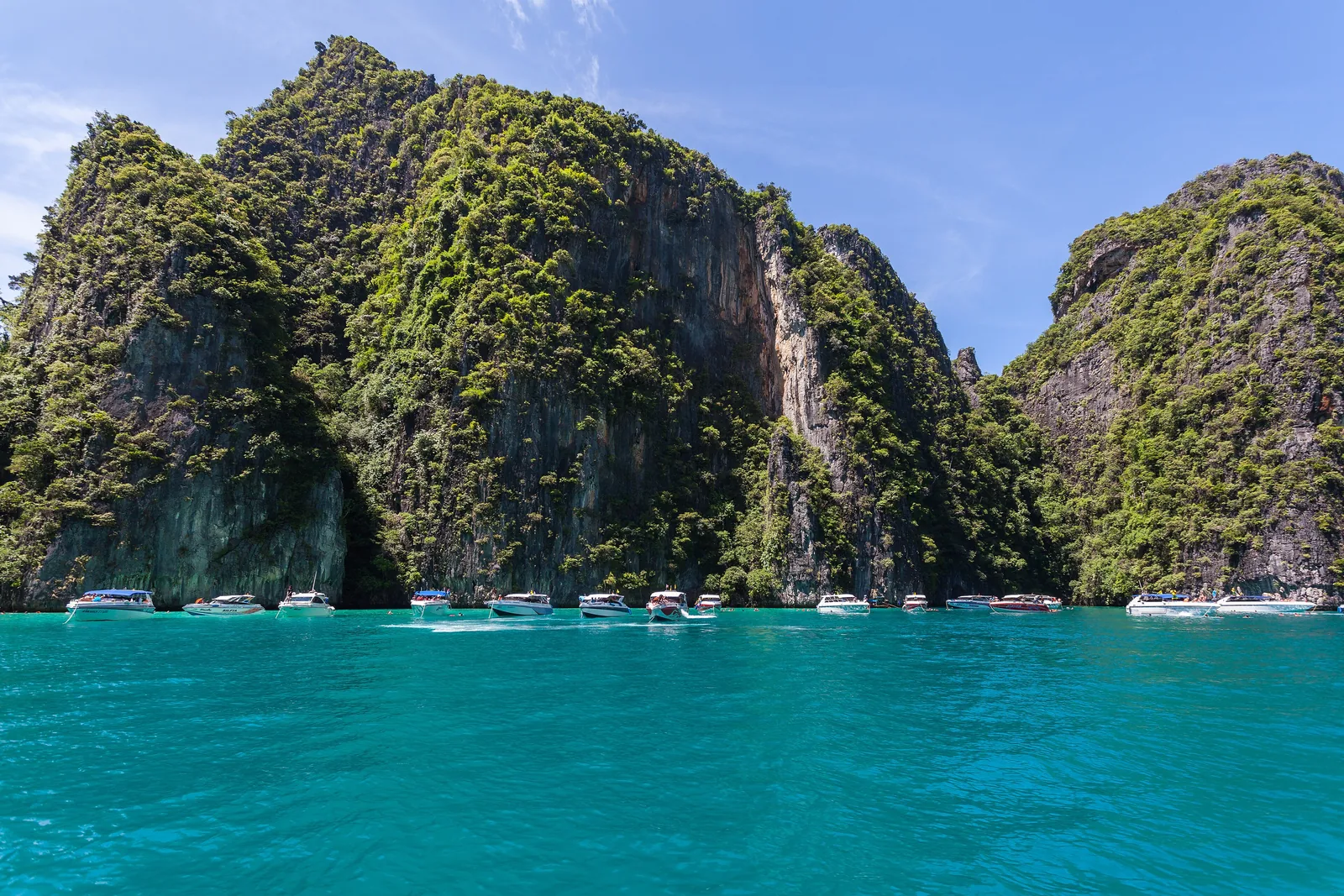 Sheer, densely overgrown limestone walls of Ko Phi Phi Le in Phi Phi National Park, with a row of speedboats on turquoise water