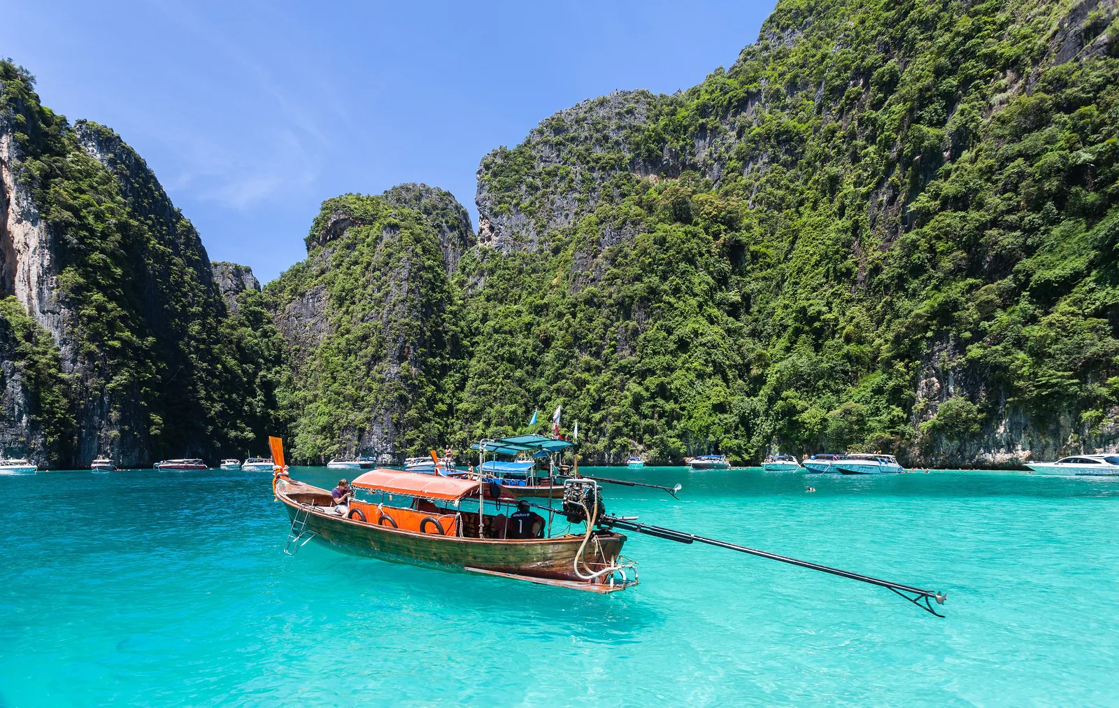 A longtail boat with an orange roof on turquoise water in front of the overgrown limestone walls of Ko Phi Phi Le