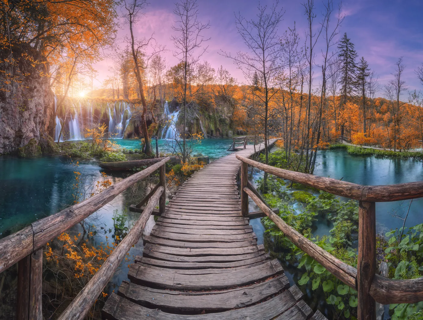 Wooden boardwalk over turquoise water in Plitvice Lakes National Park, with waterfalls and autumn forest in the evening light behind