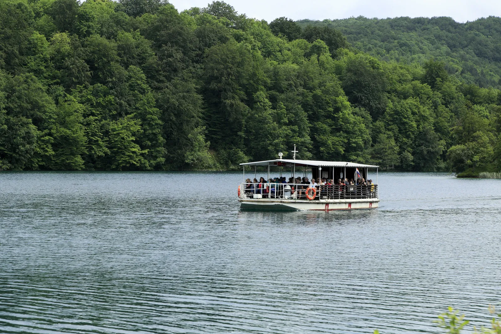 A white excursion boat full of passengers glides across the grey, shimmering Lake Kozjak in Plitvice Lakes National Park in front of a densely wooded shore