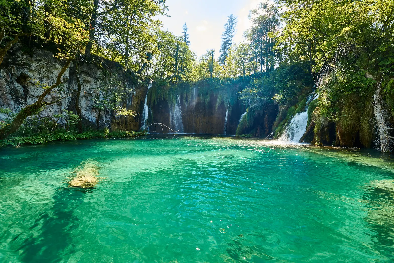 Clear turquoise water of the Upper Lakes in Plitvice Lakes National Park in front of a wooded tufa edge over which several white waterfalls of Galovački buk fall