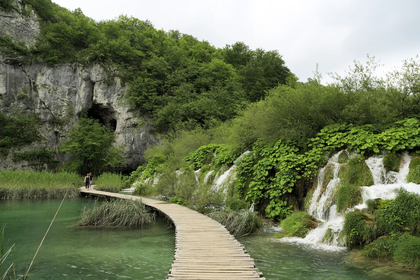 Wooden boardwalk over green water at Velike Kaskade in Plitvice Lakes National Park, with the dark opening of Šupljara cave in the pale rock face on the left