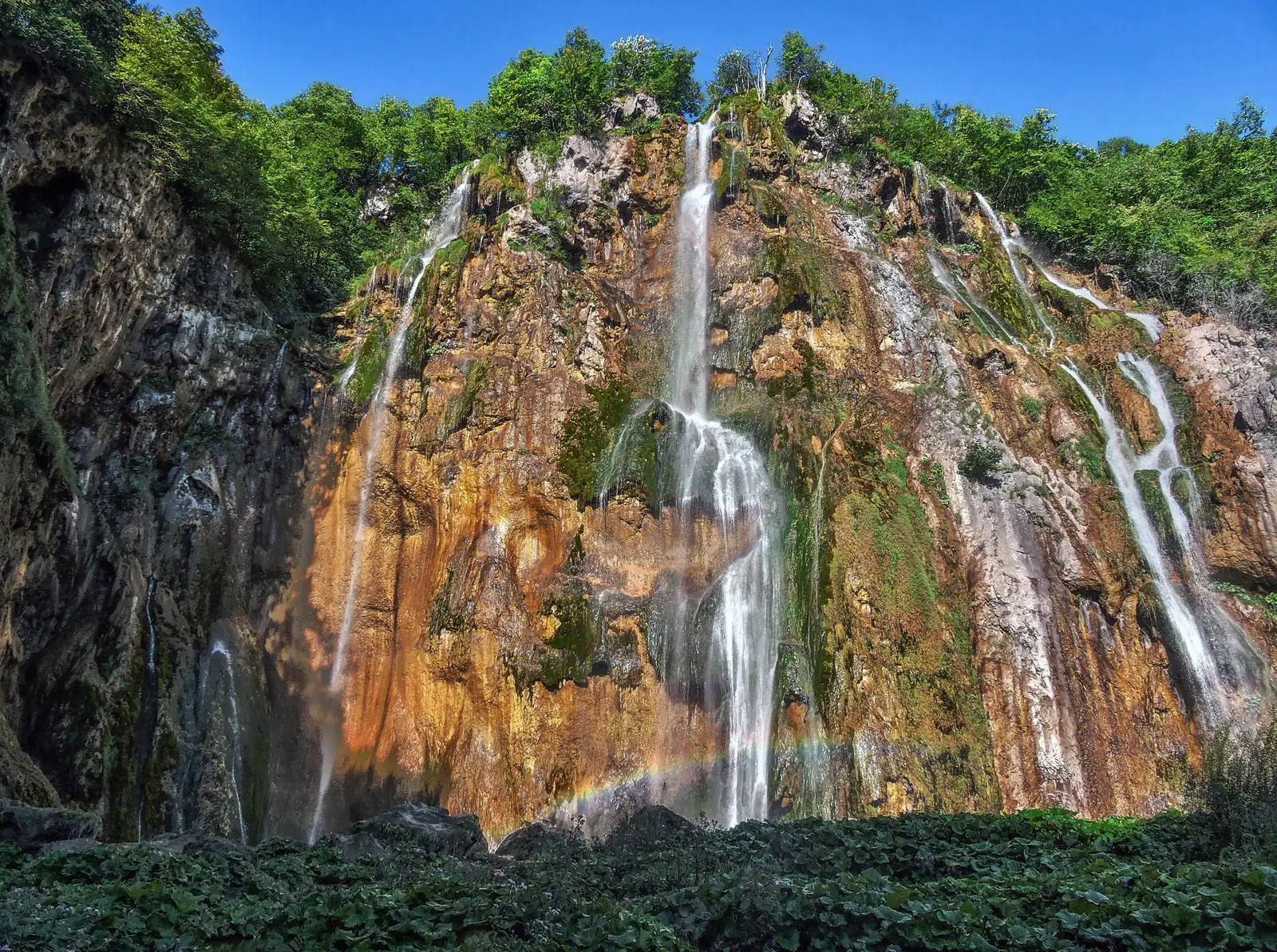 Veliki slap in Plitvice Lakes National Park falls in white strands over a rust-red, mossy rock face, with a faint rainbow in the spray below