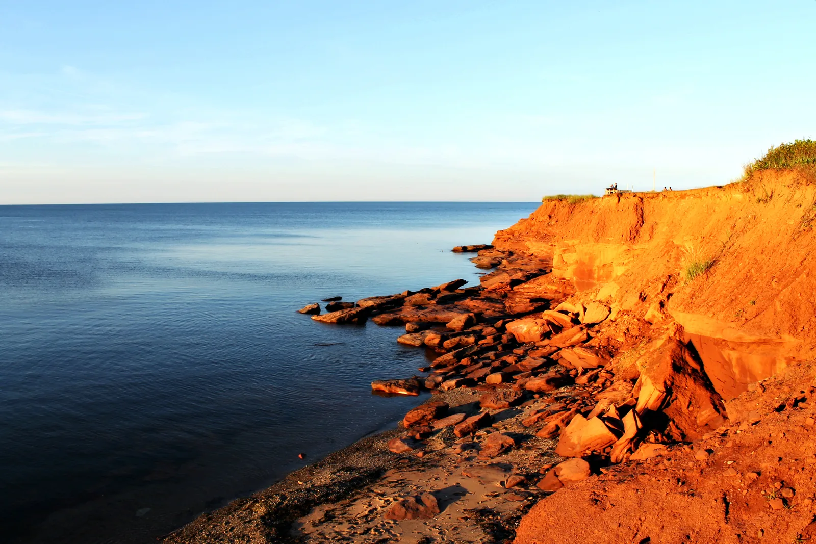 Red sandstone cliffs in Prince Edward Island National Park drop to a calm, dark blue sea in low evening light, with broken rock slabs at the foot of the cliff.