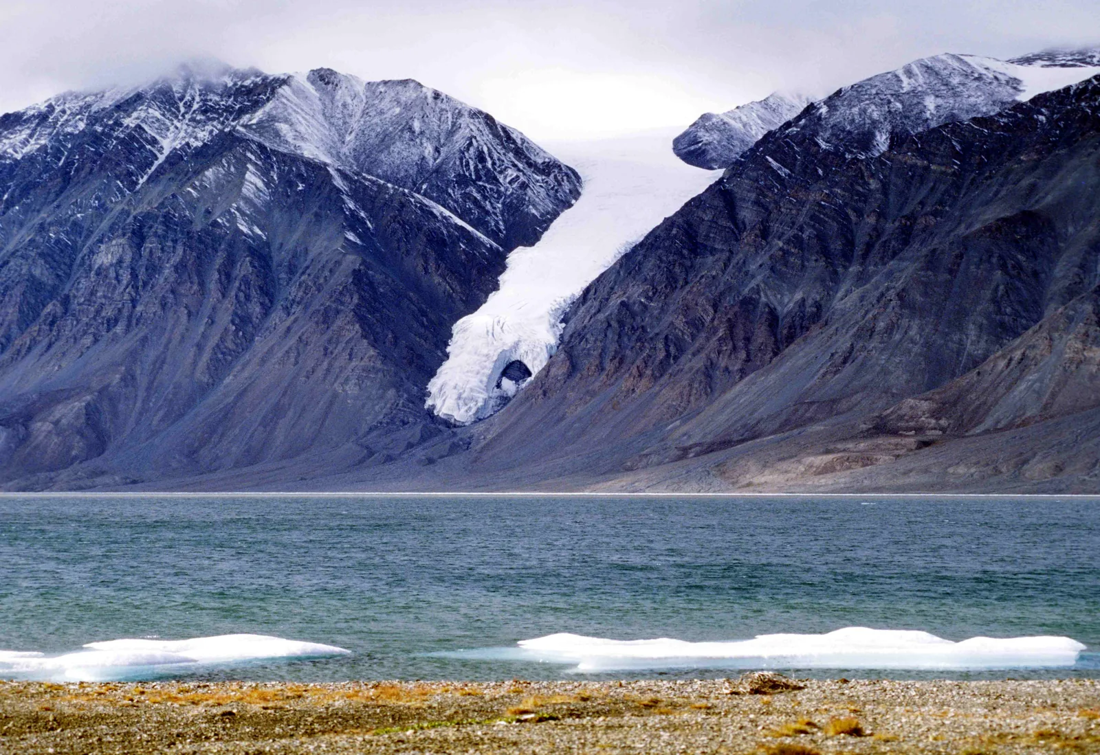 A glacier in Quttinirpaaq National Park pushes down between dark, snow-streaked mountains to the shore of a fiord, with ice floes on the beach