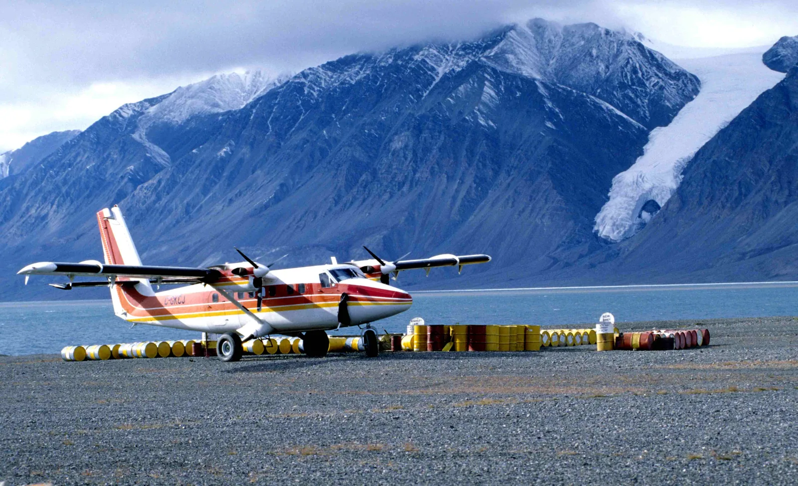A red and white Twin Otter on a gravel airstrip between rows of fuel drums in Quttinirpaaq National Park, a fiord and glaciated mountains behind