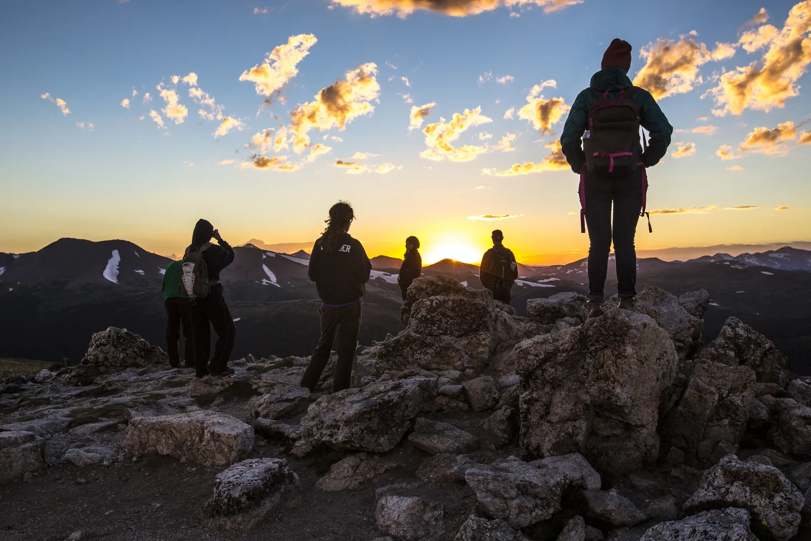 Several people stand as silhouettes on boulders in Rocky Mountain National Park, looking across the tundra at an orange sunset behind mountain ranges
