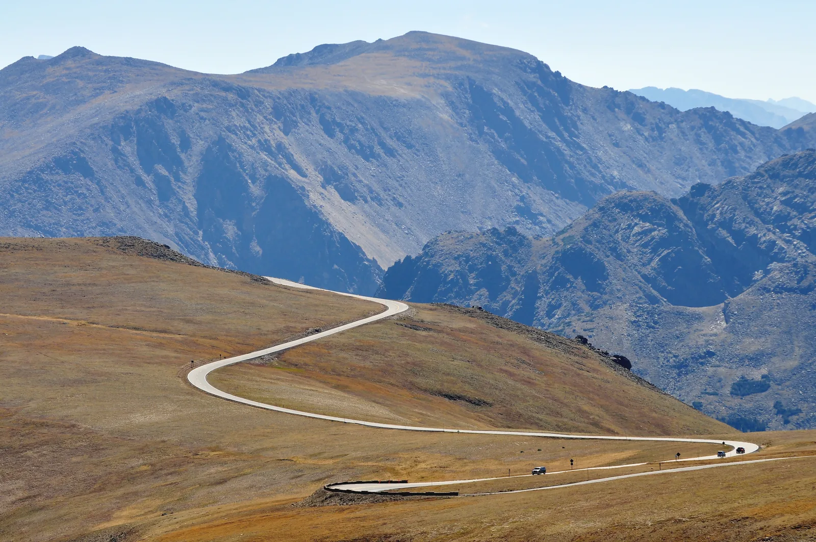 A narrow paved road in Rocky Mountain National Park sweeps in wide curves across treeless, golden-brown uplands, with tiny cars on it and bare rocky peaks behind