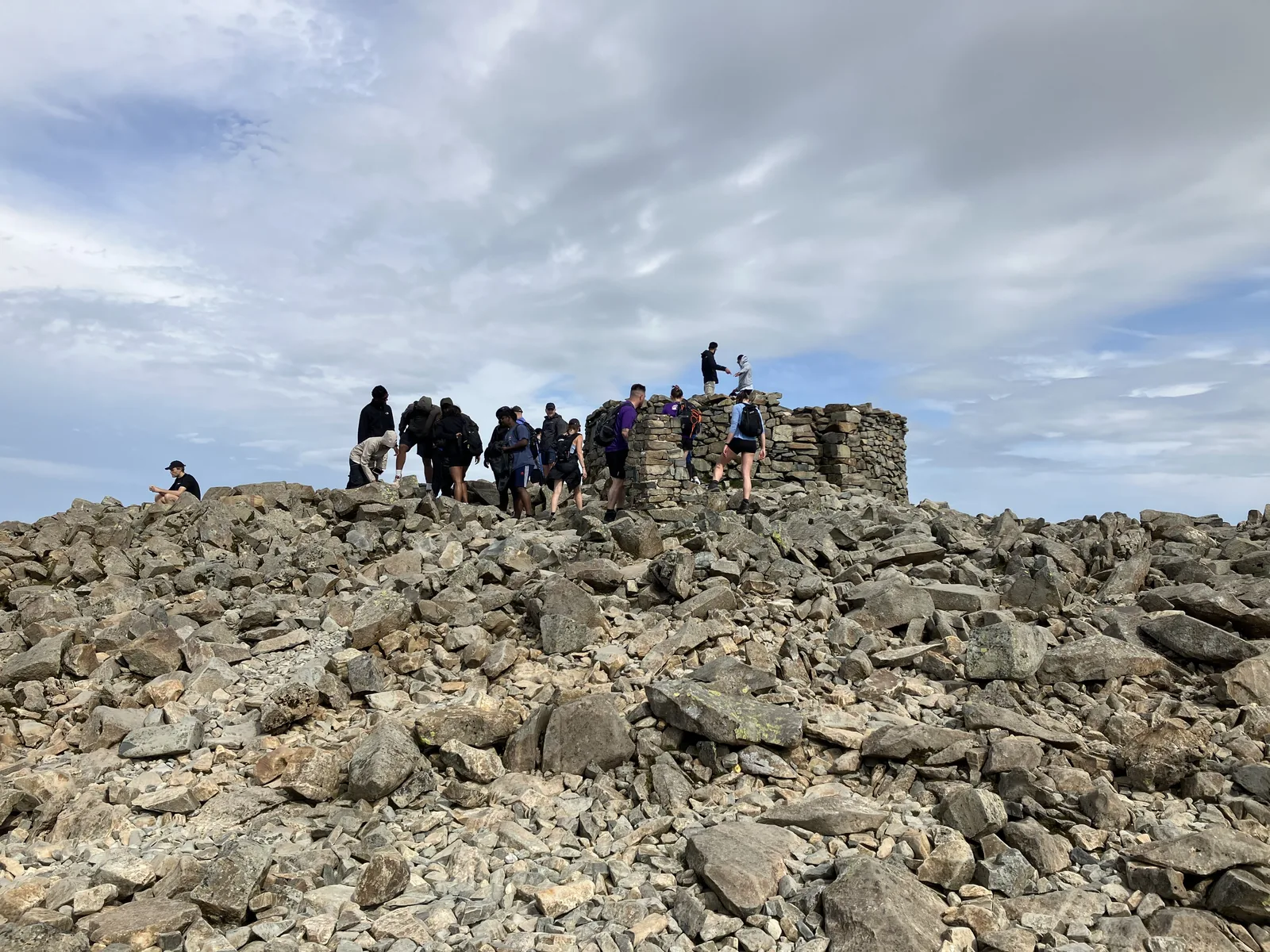 A group of walkers at the round summit cairn of Scafell Pike in the Lake District National Park, with scree all around